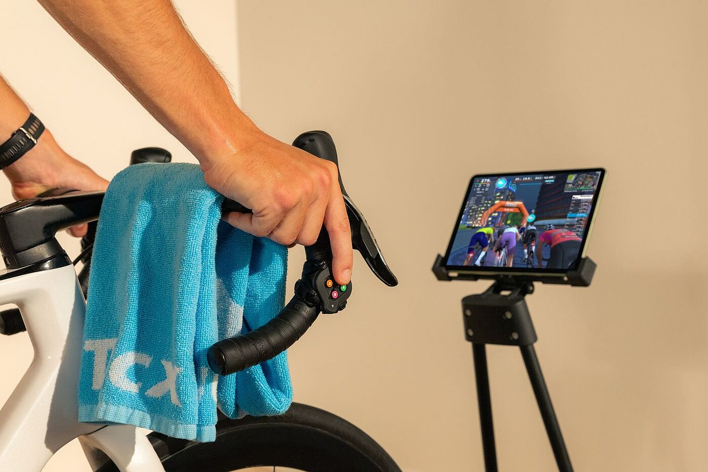 Eine Hand hält den Lenker eines Rennrads. Rechts steht ein Tablet auf einem Ständer, auf dem ein Radrennen am Bildschirm zu sehen ist.