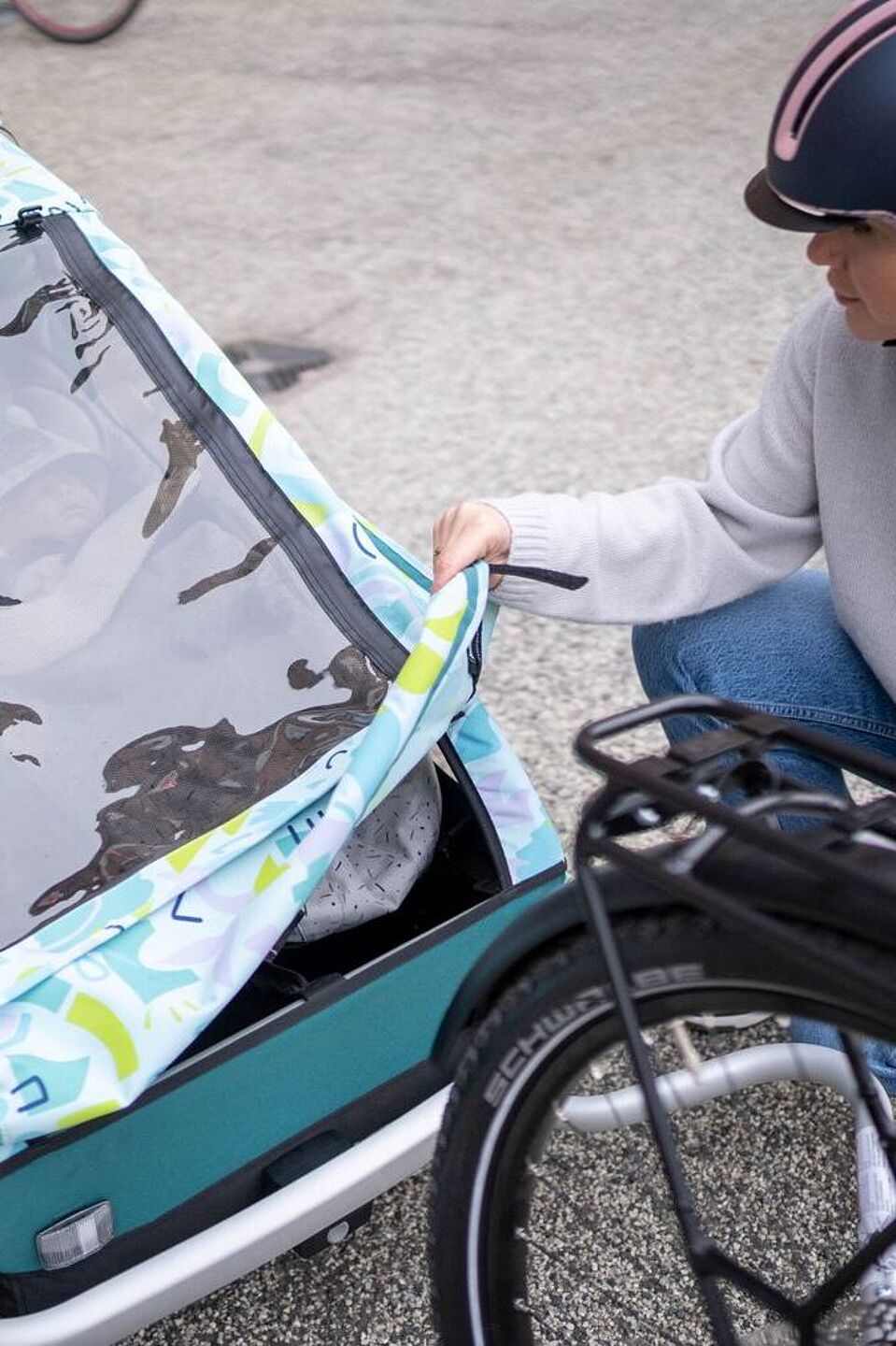 Ein Regenverdeck schützt Kinder im Anhänger vor Regen und Schnee sowie vor Schmutz und aufgewirbelten Steinchen vom Hinterrad des Zugfahrrads. Eine Frau mit Fahrradhelm kniet vor Fahrradanhänger für Kinder, der an ihrem Fahrrad befestigt ist und schließt das Regenverdeck, um ihr Kind im Anhänger zu schützen.